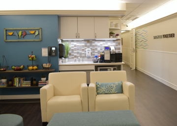 A hospital's Ronald McDonald Family Room common area, two beige armchairs and coffee table in foreground and kitchen, fresh fruit, wall art, and hospital phone in background