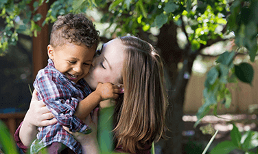 An RMHC family, mom Rachel Jolivard holding son Jacob by the waist and kissing his cheek, both enjoying the sun