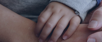 Link background, extreme close-up on the hands of an infant and parents in embrace, a small hand holding onto one parent's who is holding the hand of another parent