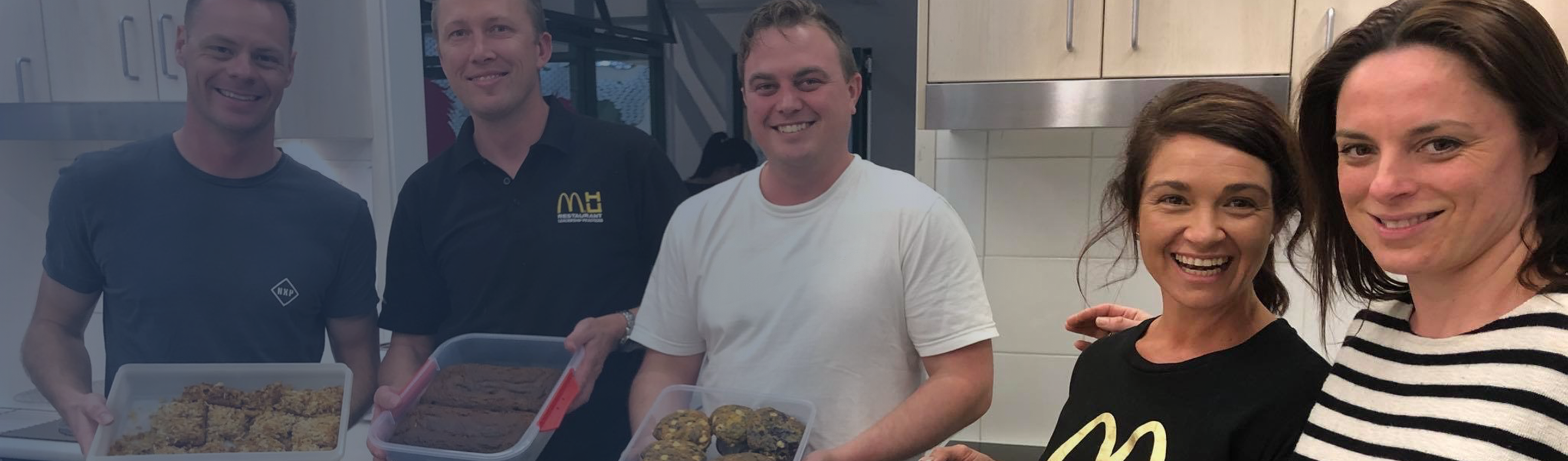 McDonald's volunteers smiling, posed holding prepared dishes, a female pair in foreground focus on the right, a male trio in soft focus on the left