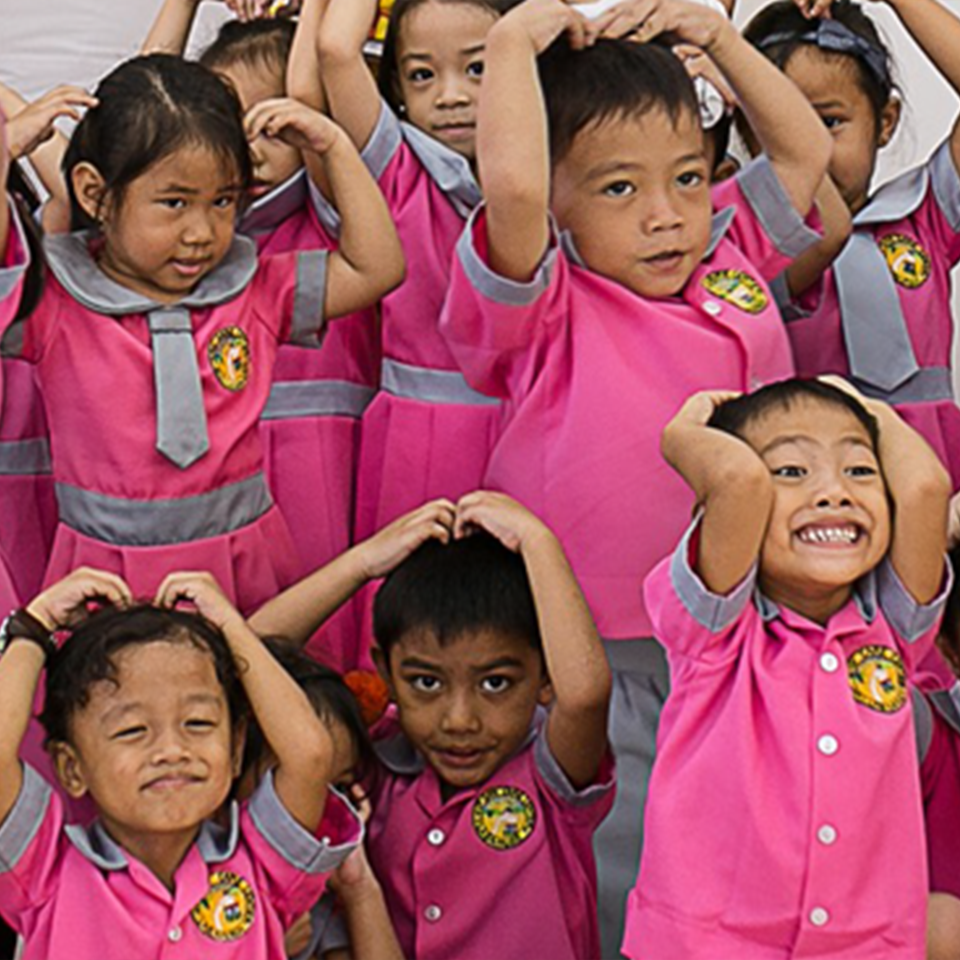 Smiling children at a Bahay Bulilit Learning Center, wearing matching pink school uniforms, all posed with hands on heads