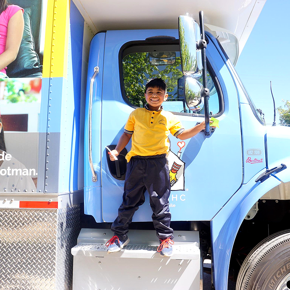 Sun shining on a smiling boy standing in the shade of the passenger-side mirror, his heels balancing on the step up to the Care Mobile cab