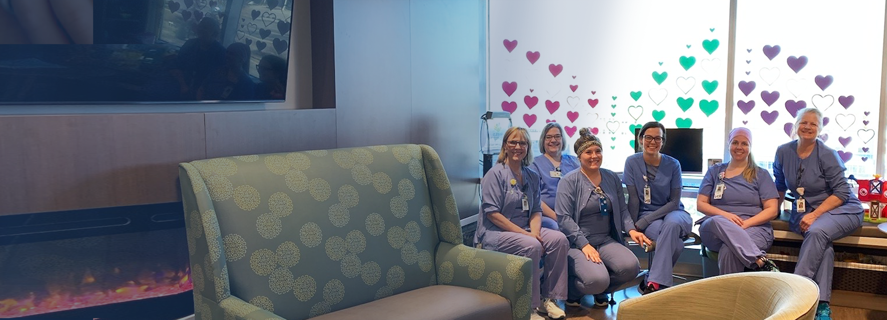 Pediatric nurses sit for the camera, lit by the window behind them decorated with multiple colors of heart symbols, furniture in soft focus in front