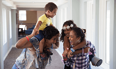 RMHC family, Vasco Gonzalez and mom Antuane playing, a boy carried sitting on one woman's shoulder and a girl carried on the back of another